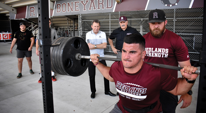 Linebacker Nick Castro ’19 (front) prepares to lift 545 pounds, spotted by Strength and Conditioning Coach and Fitness Center Director Joe Buckley (right), as President Ralph Kuncl (left) and Coach Mike Maynard look on. (Photo by Carlos Puma)